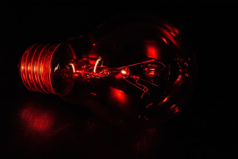 Red glowing light bulb with a visible filament and metallic base set against a dark, pitch-black background.