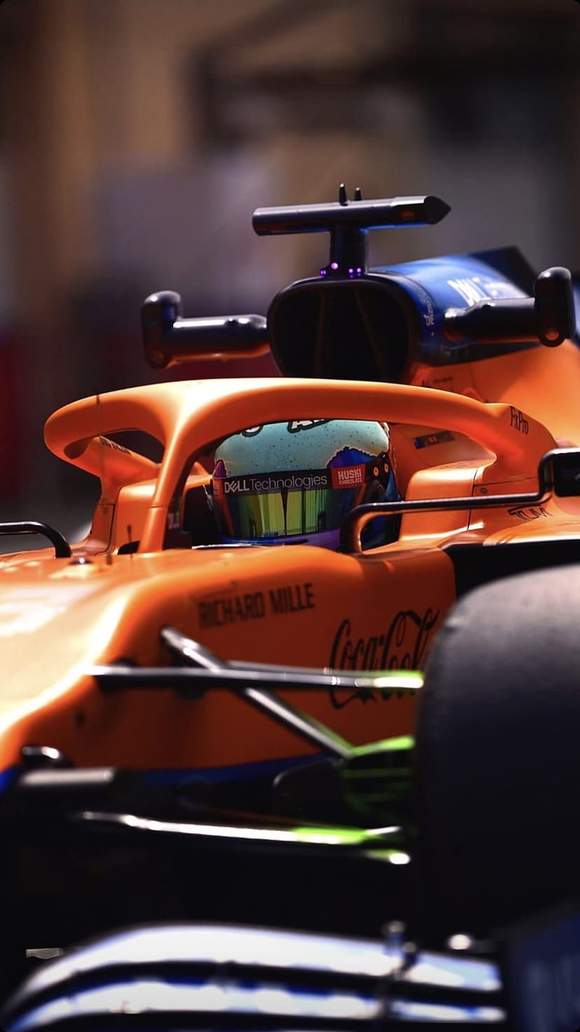 Formula 1 driver in an orange and blue cockpit wears a racing helmet featuring prominent corporate branding.
