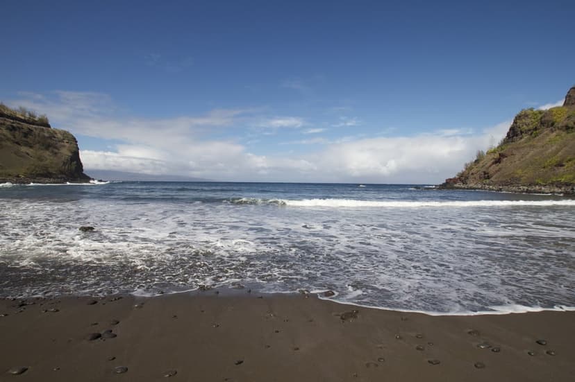 Black sand beach curves toward steep green cliffs under a blue sky with white surf washing onto the dark shoreline.