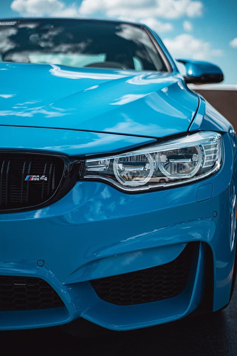Blue BMW M4 headlight and kidney grille with a metallic finish reflecting a cloudy sky in a vertical close-up.