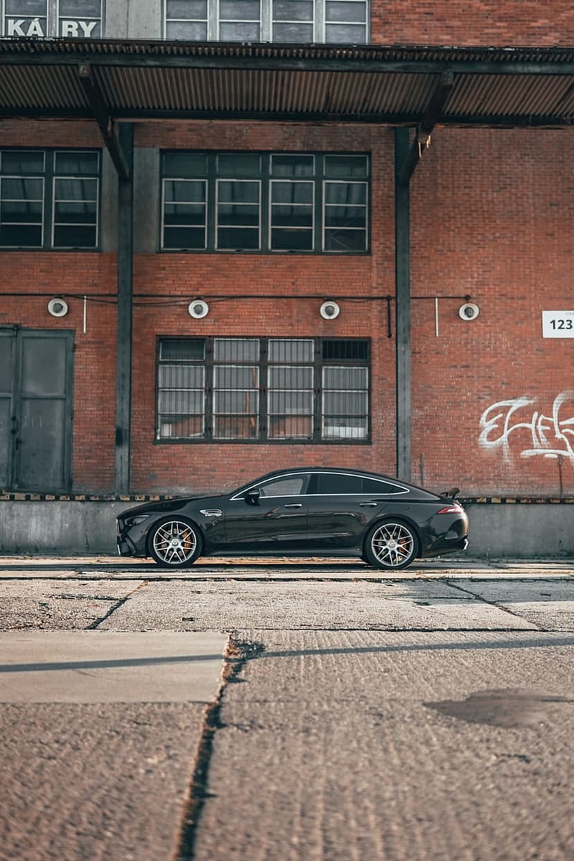 Dark AMG GT 4-Door Coupe parked on cracked concrete beside a brick industrial building with graffiti.
