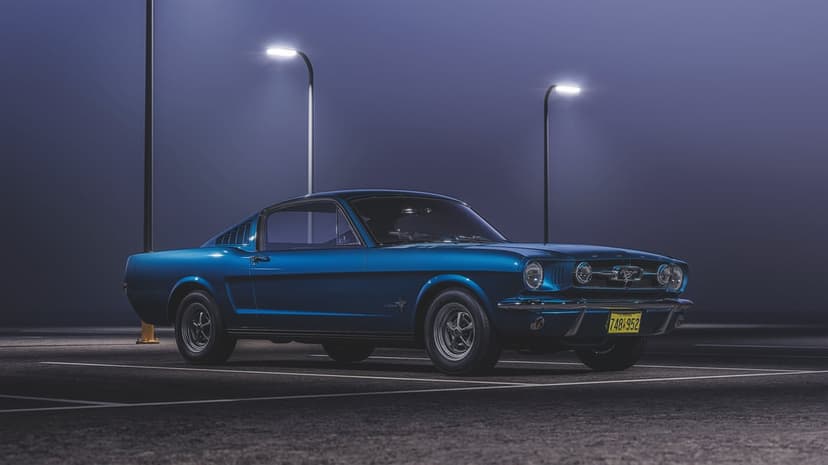 Classic blue Ford Mustang Fastback parked in a misty parking lot under two glowing streetlights at night.