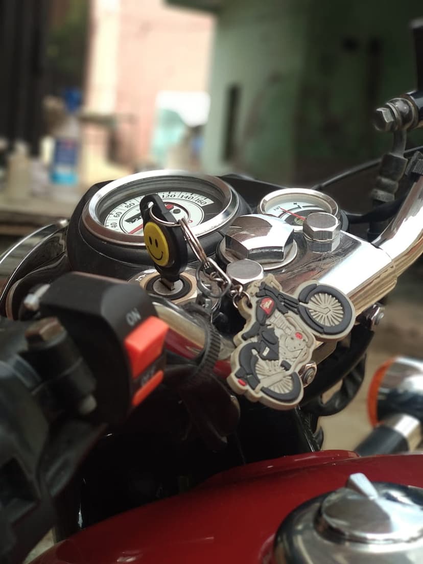 Motorcycle handlebar features a speedometer, ignition key with a smiley face keychain, and a red fuel tank.