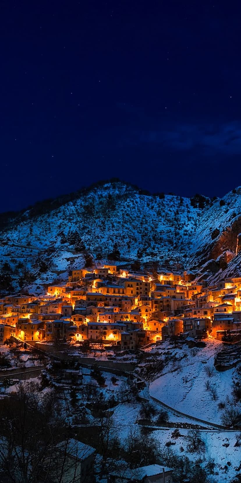 Castelmezzano village lights glow against snow-covered mountains under a dark night sky with visible stars.