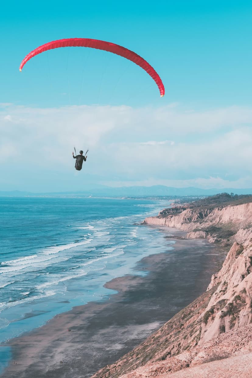 A red paraglider flies high above steep coastal cliffs and turquoise ocean waves under a clear blue sky.