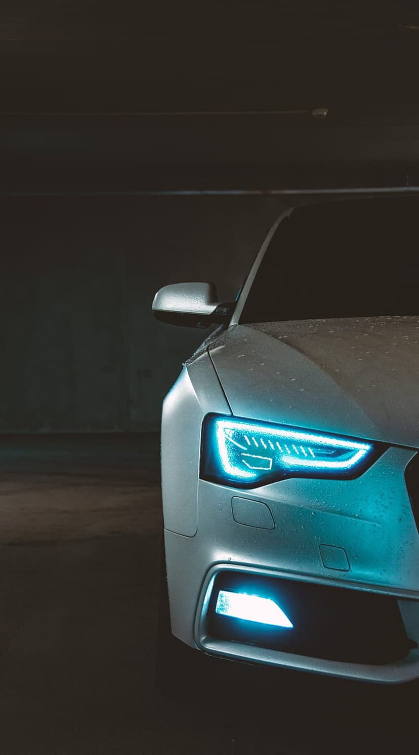Silver Audi A5 front headlight glowing with blue LED light on a wet metallic surface against a dark background.