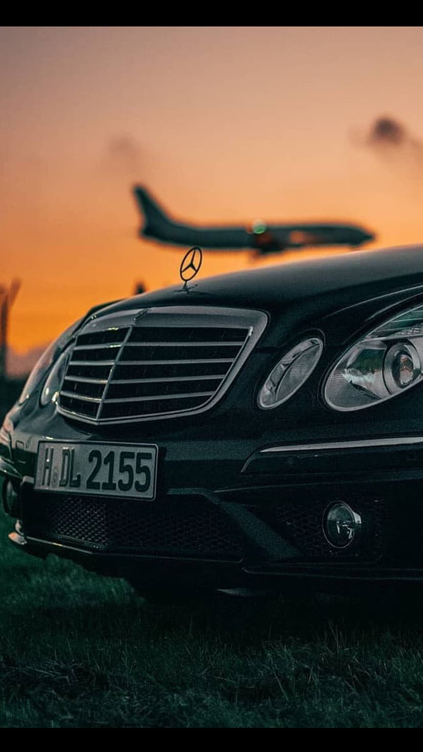 Black Mercedes-Benz on grass with a blurred airplane taking off against a bright orange sunset sky.