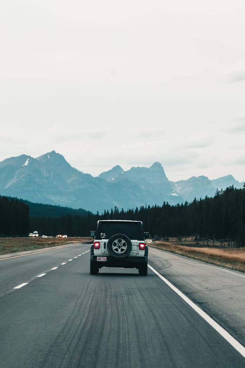 White Jeep Wrangler driving on a paved highway toward distant mountain peaks under a grey overcast sky.