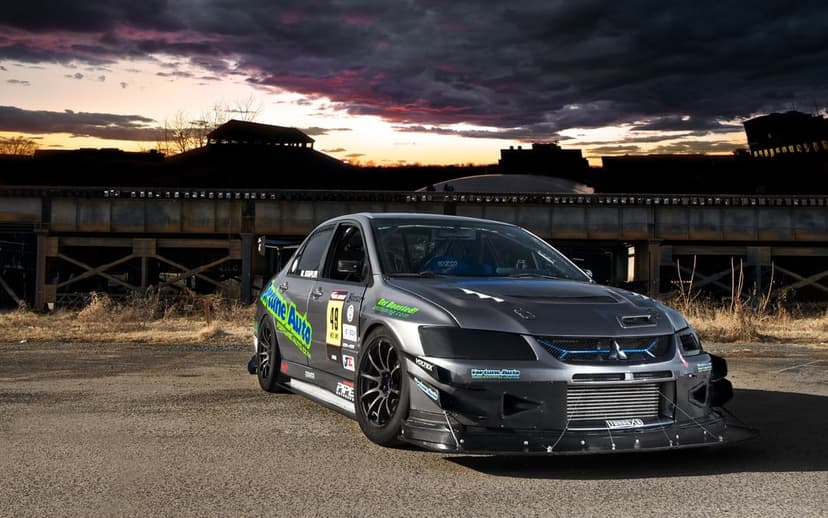 Grey modified Mitsubishi Evo with track aerodynamics parked before an industrial bridge under a purple sunset sky.