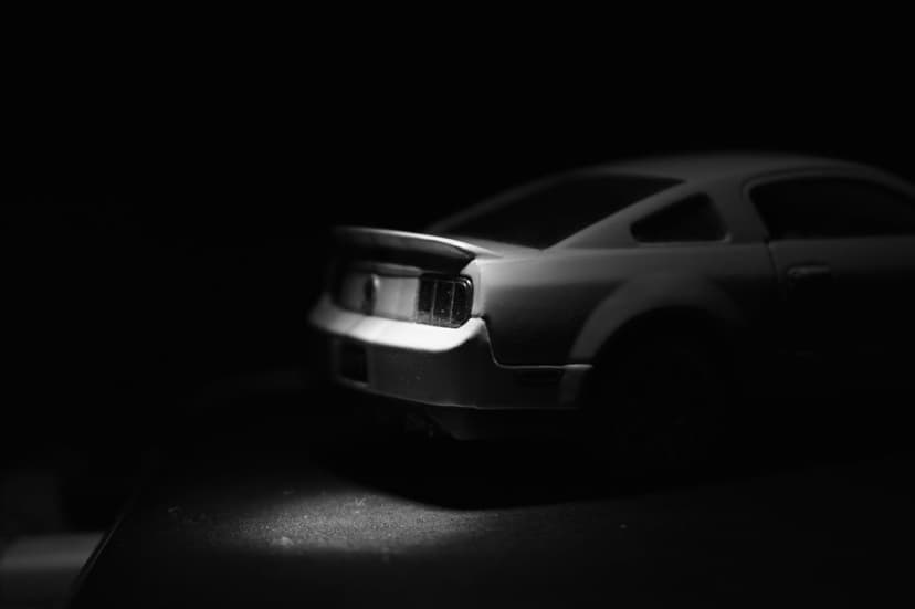 Ford Mustang toy car rear end with illuminated taillights emerging from a dark black background in monochrome.