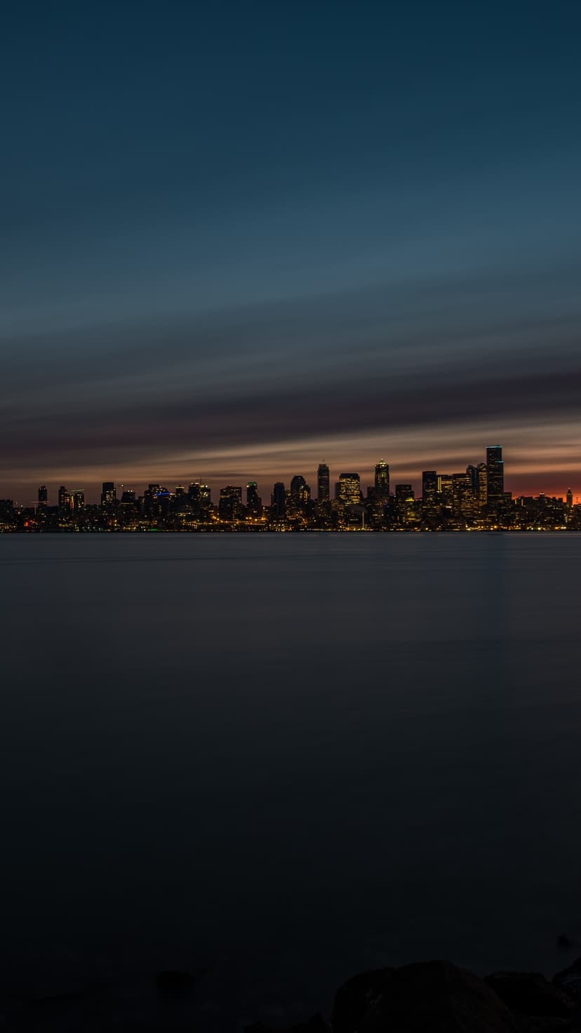Luminous city skyscrapers overlook a dark harbor under a fading orange horizon during late evening twilight.