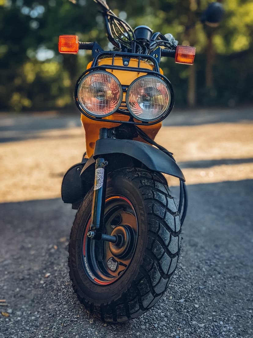 Yellow retro scooter with dual round headlights and orange signals stands on a textured gravel driveway.