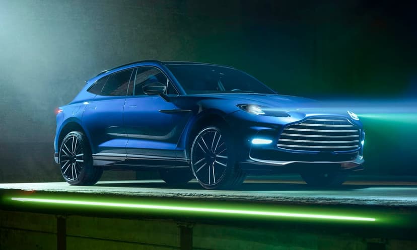 Blue Aston Martin DBX707 luxury SUV on a platform with green neon lights and glowing headlights in a dark room.