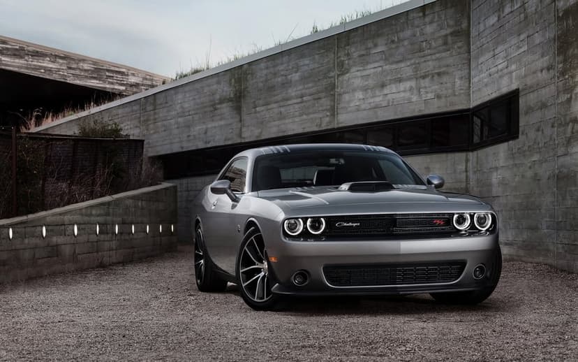 Silver Dodge Challenger with glowing halo headlights parked on gravel beside a modern concrete wall at twilight.