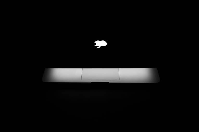 Apple logo glowing on a black MacBook keyboard with visible keys and trackpad in a dark minimalist setting.