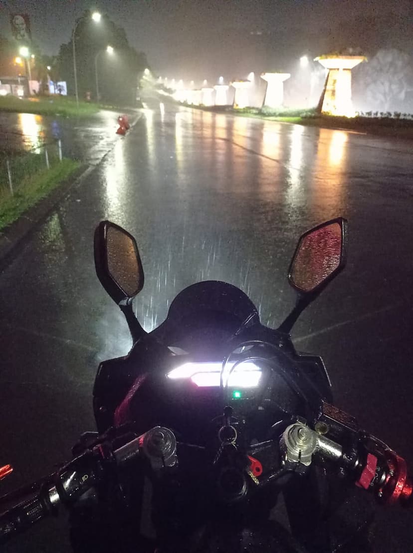 Motorcycle handlebars and glowing dashboard on a wet asphalt road at night with raindrops and blurred city lights.
