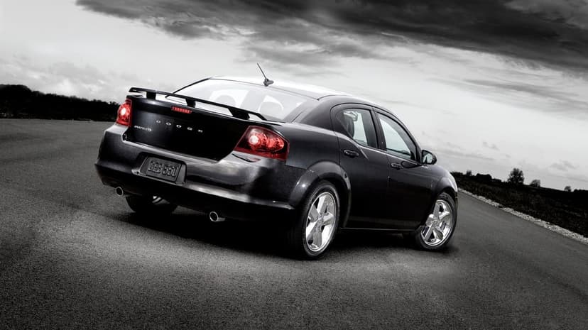 Rear view of a black Dodge Avenger sedan on an asphalt road under a dramatic, cloudy sky with red tail lights.