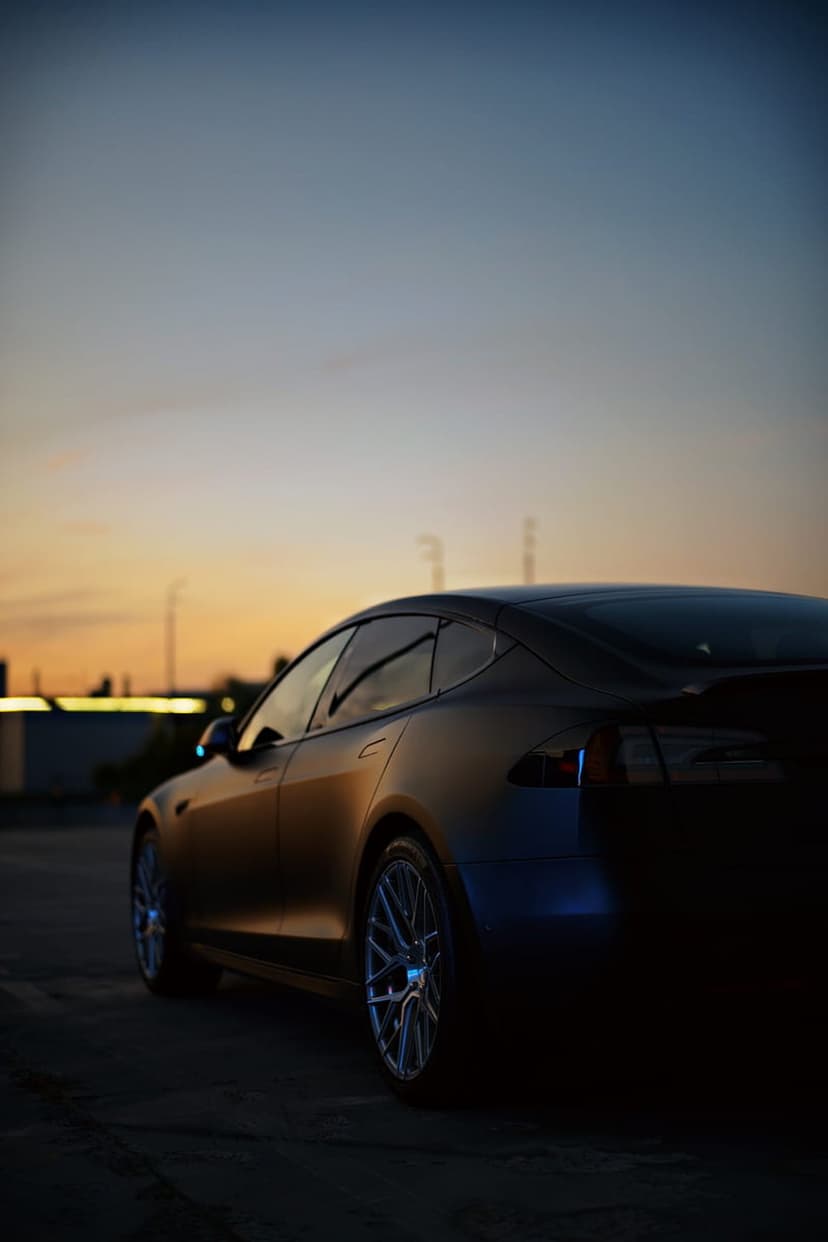 Tesla Model S rear view at dusk with orange sunset highlights on its frame against a silhouetted industrial sky.