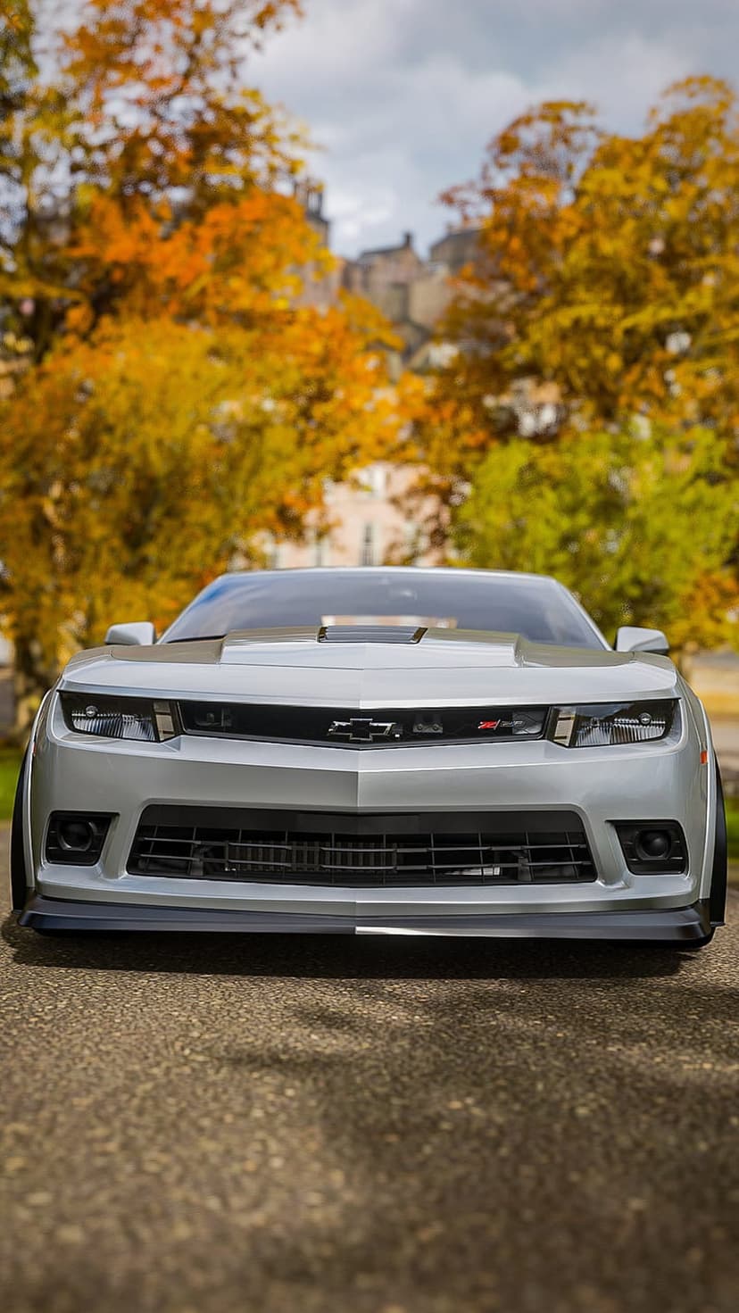 Silver Chevrolet Camaro ZL1 parked on a paved road surrounded by orange and yellow autumn trees in a forest.