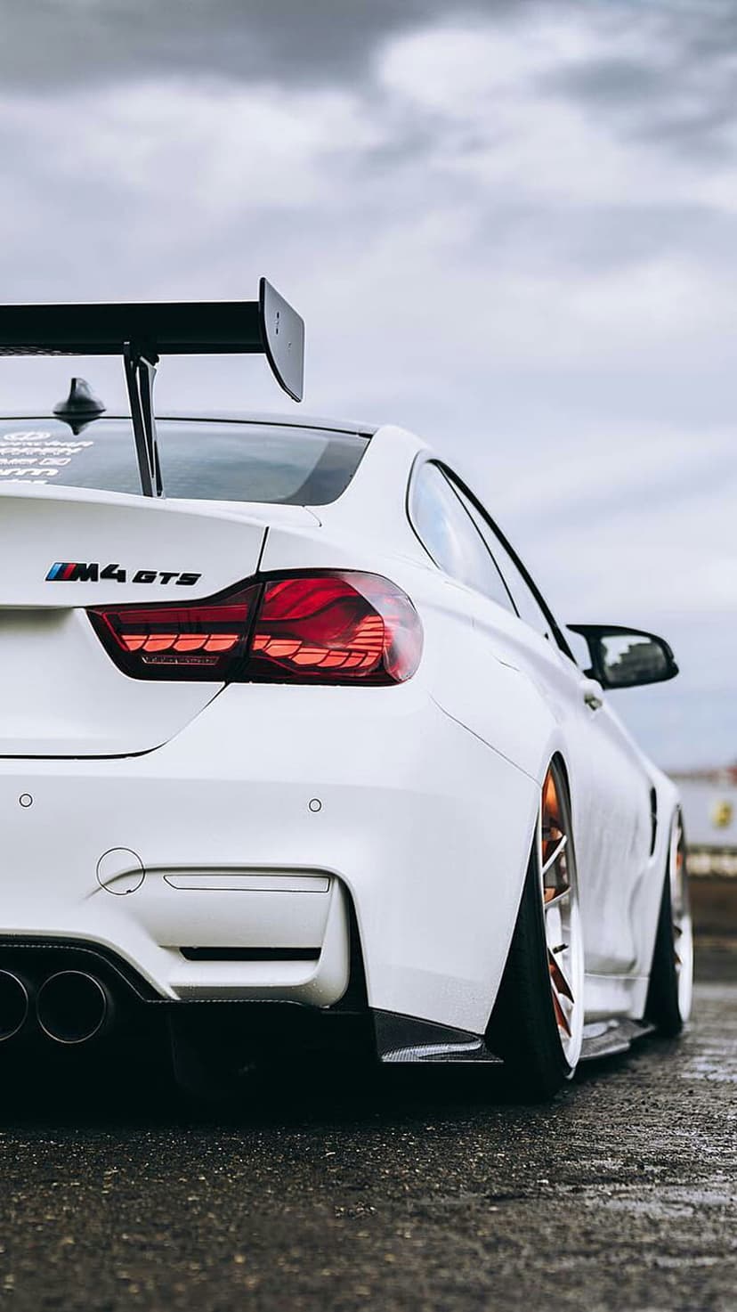 BMW M4 GTS rear view in white with a large black spoiler, glowing red taillights, and a cloudy sky background.