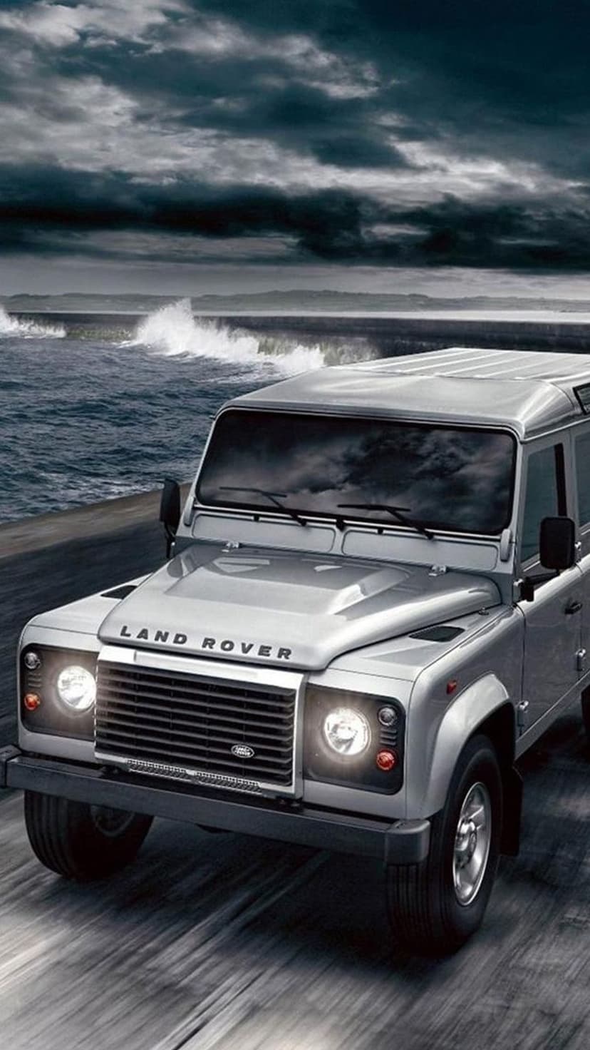 Silver Land Rover Defender with glowing headlights driving along a sea wall under a dark stormy sky.