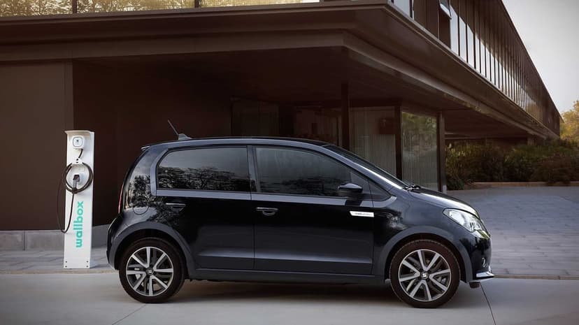 Black SEAT Mii Electric car parked next to a white wallbox charger in front of a modern glass building.