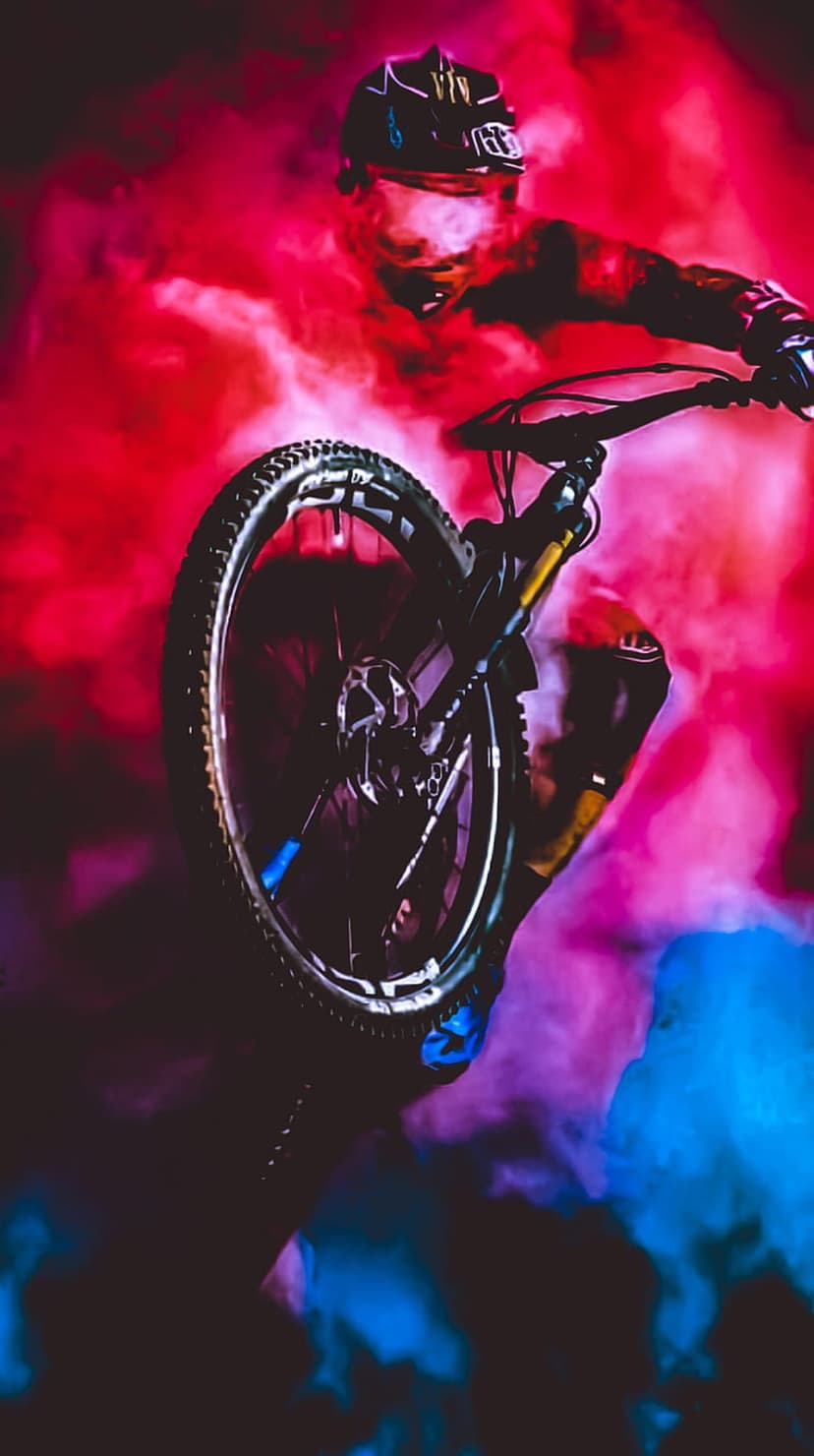 Mountain biker jumps mid-air through swirling red and blue smoke trails wearing a full-face helmet and safety gear.