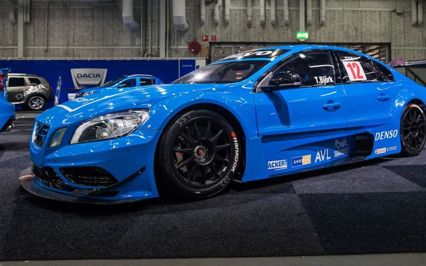 Blue Volvo racing car with wide body kit and black wheels on a dark showroom floor in a side profile view.