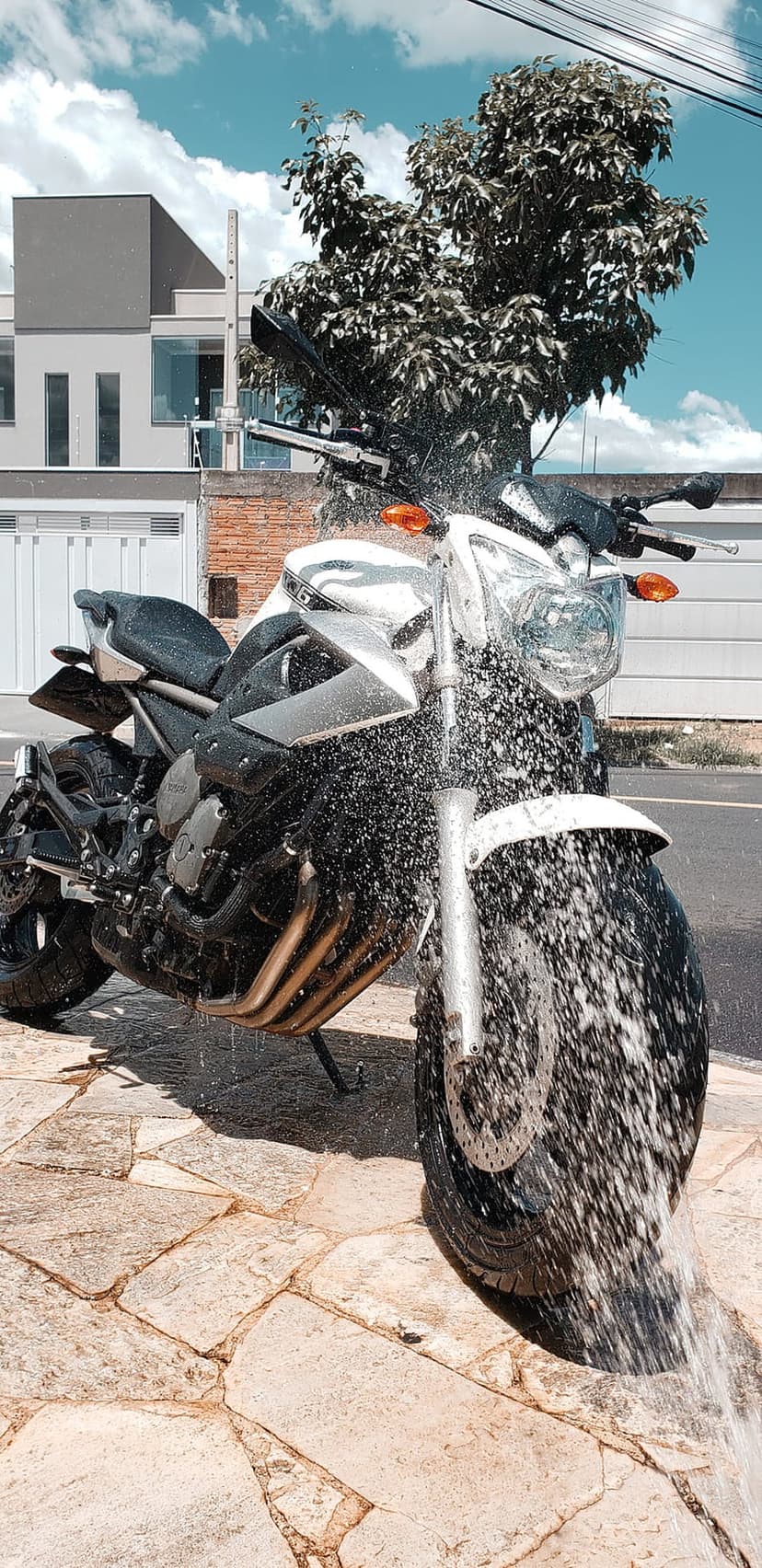 Motorcycle front tire and handlebars glistening under a stream of high-pressure water spray in a garden setting.