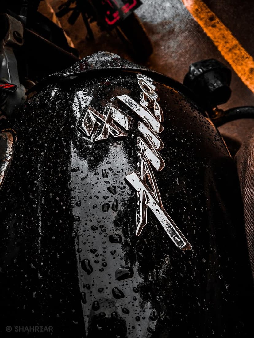 Black motorcycle fuel tank with a silver chrome emblem covered in small, clear raindrops after a rain shower.