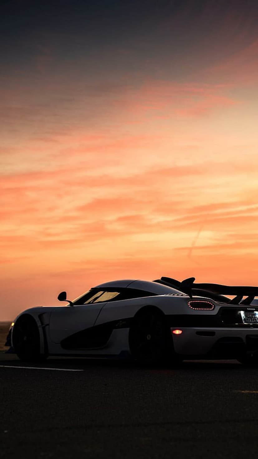Koenigsegg hypercar silhouette with a large rear wing against a glowing orange and pink sunset sky at low angle.