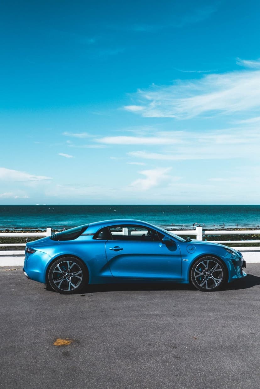 Metallic blue Alpine A110 sports car parked on a coastal road overlooking the ocean under a clear bright sky.