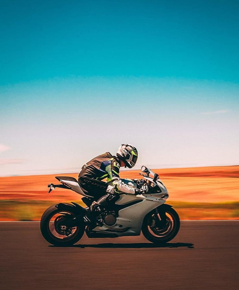 Motorcyclist in a white racing suit leans a sportbike into a corner on an asphalt track under a blue sky.