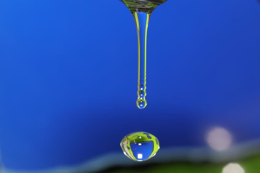 Crystal water droplet falling from a metal tap reflecting a green forest and blue sky in a sharp macro shot.