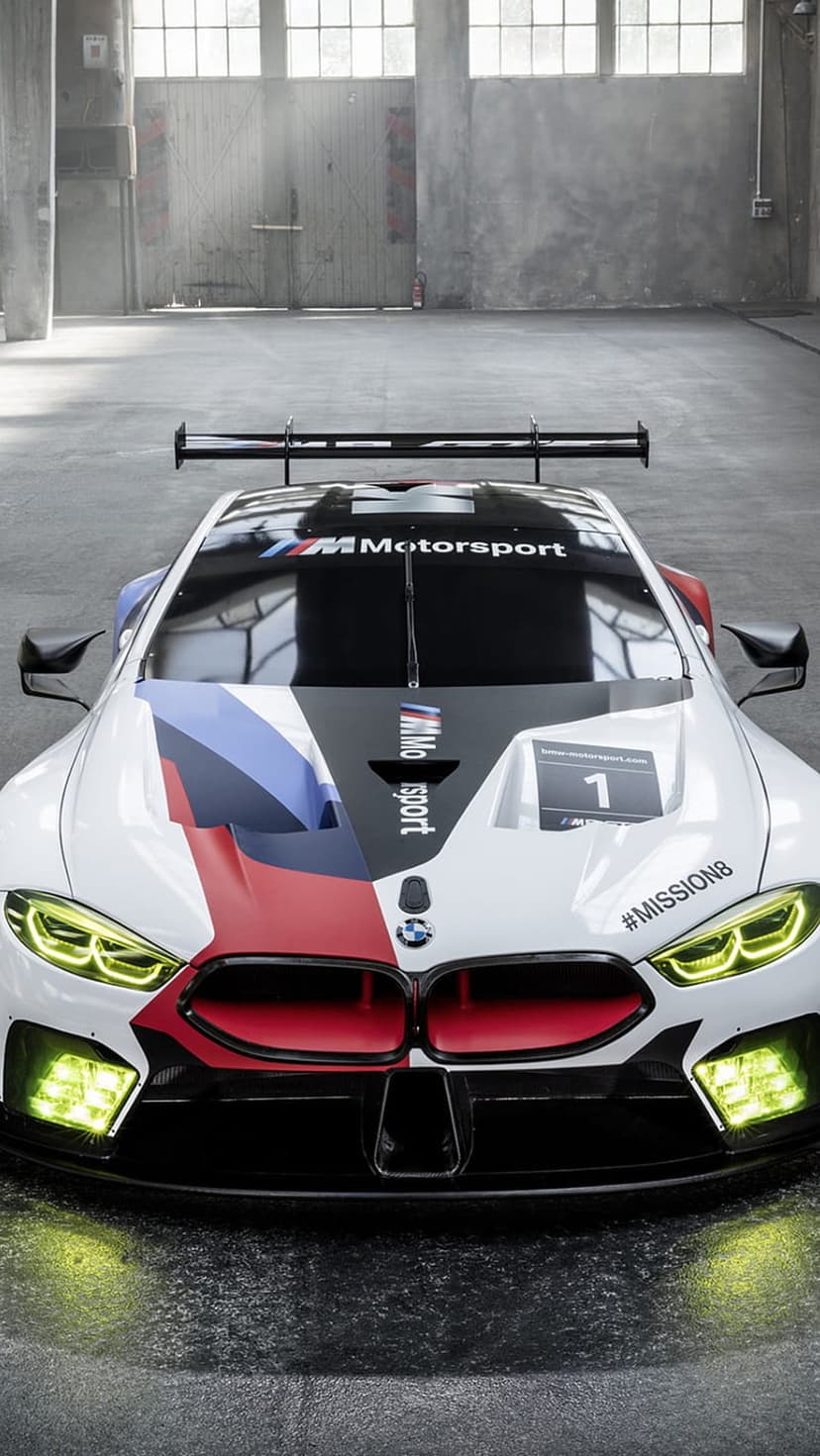 BMW M8 GTE race car with glowing yellow headlights and M stripes parked on a reflective concrete warehouse floor.