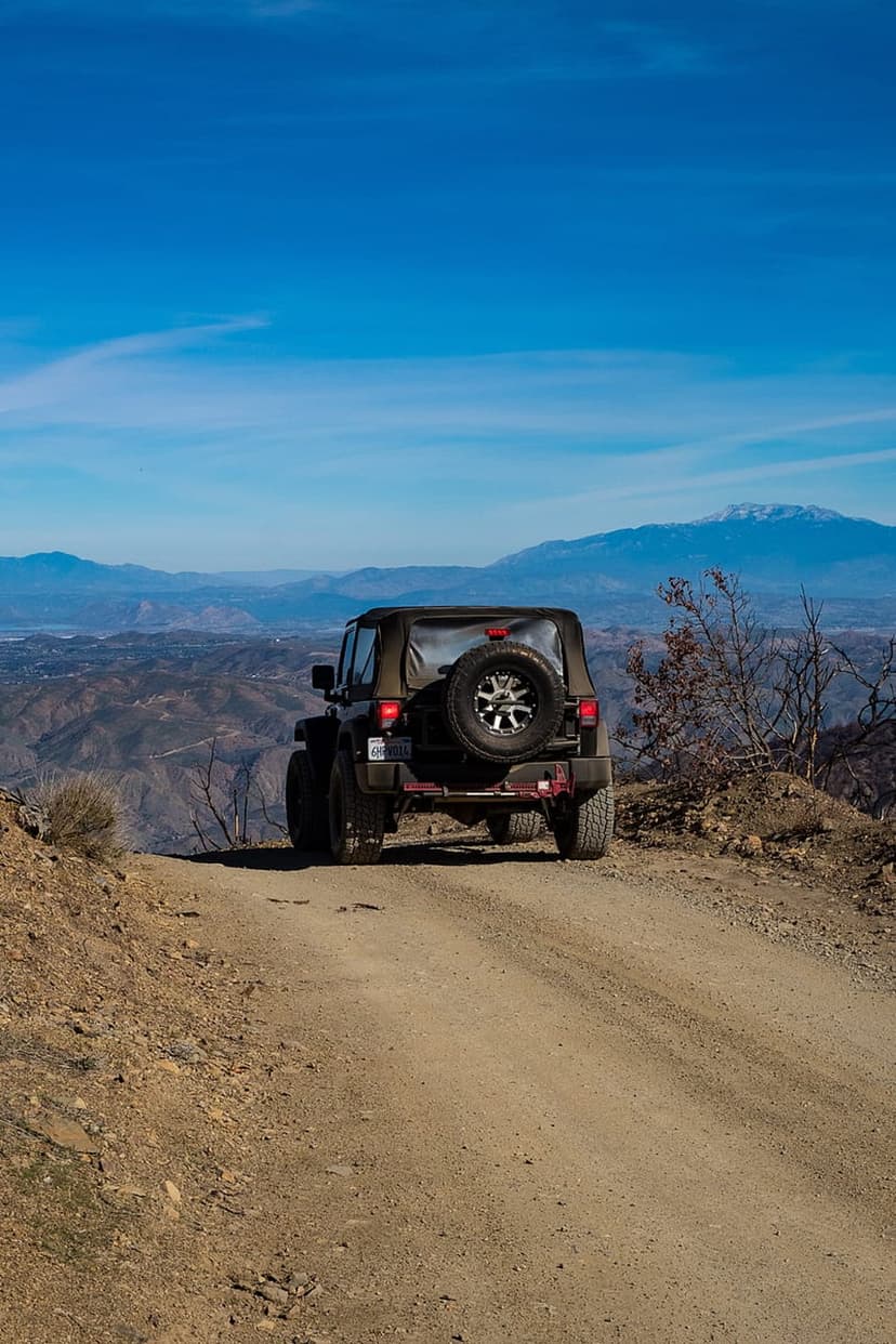 Jeep Wrangler drives on a dusty mountain road toward snow-capped peaks under a clear blue sky in vertical view.