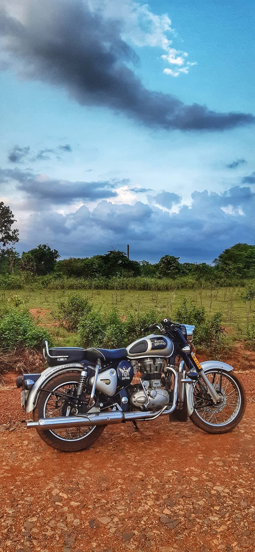 Silver Royal Enfield motorcycle on a red dirt path with green fields and dark storm clouds in the background.