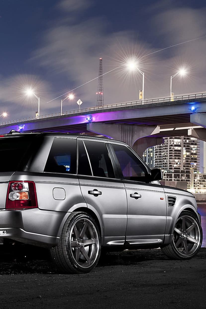 Silver Range Rover Sport parked on dark pavement under a bridge with purple neon lights and dramatic night sky.