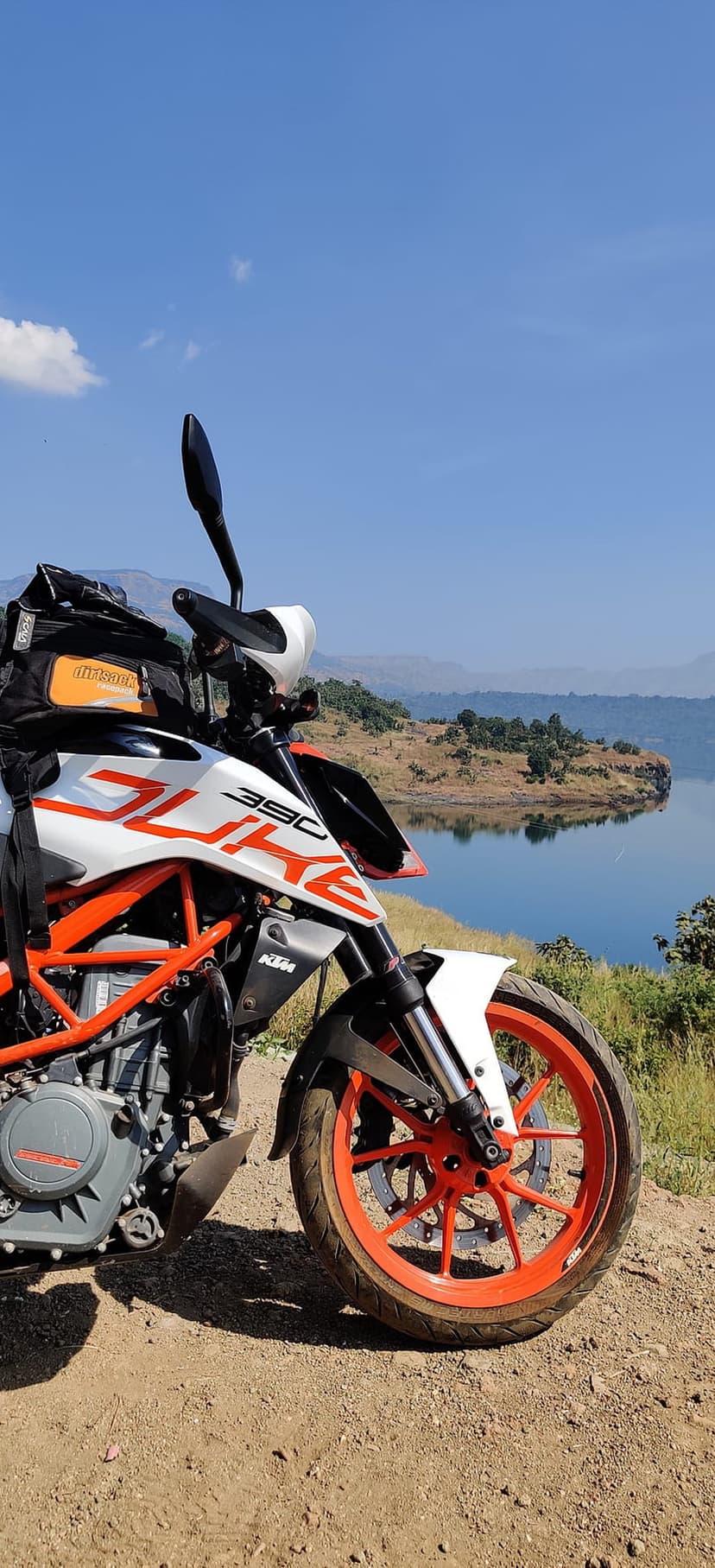 KTM Duke motorcycle with orange wheels parked on a dirt path facing a calm blue lake and distant green mountains.