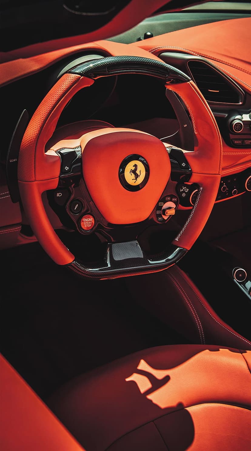 Red leather Ferrari steering wheel with carbon fiber accents and the yellow Prancing Horse emblem in a car cockpit.