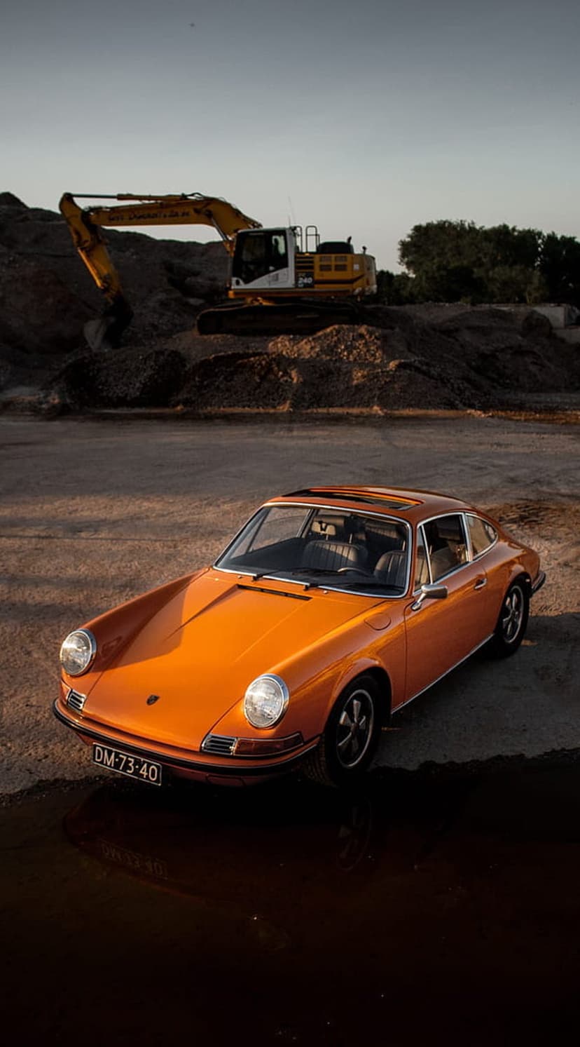 Orange Porsche 911 parked on a dirt lot reflects in a puddle near a large yellow excavator during a soft sunset.