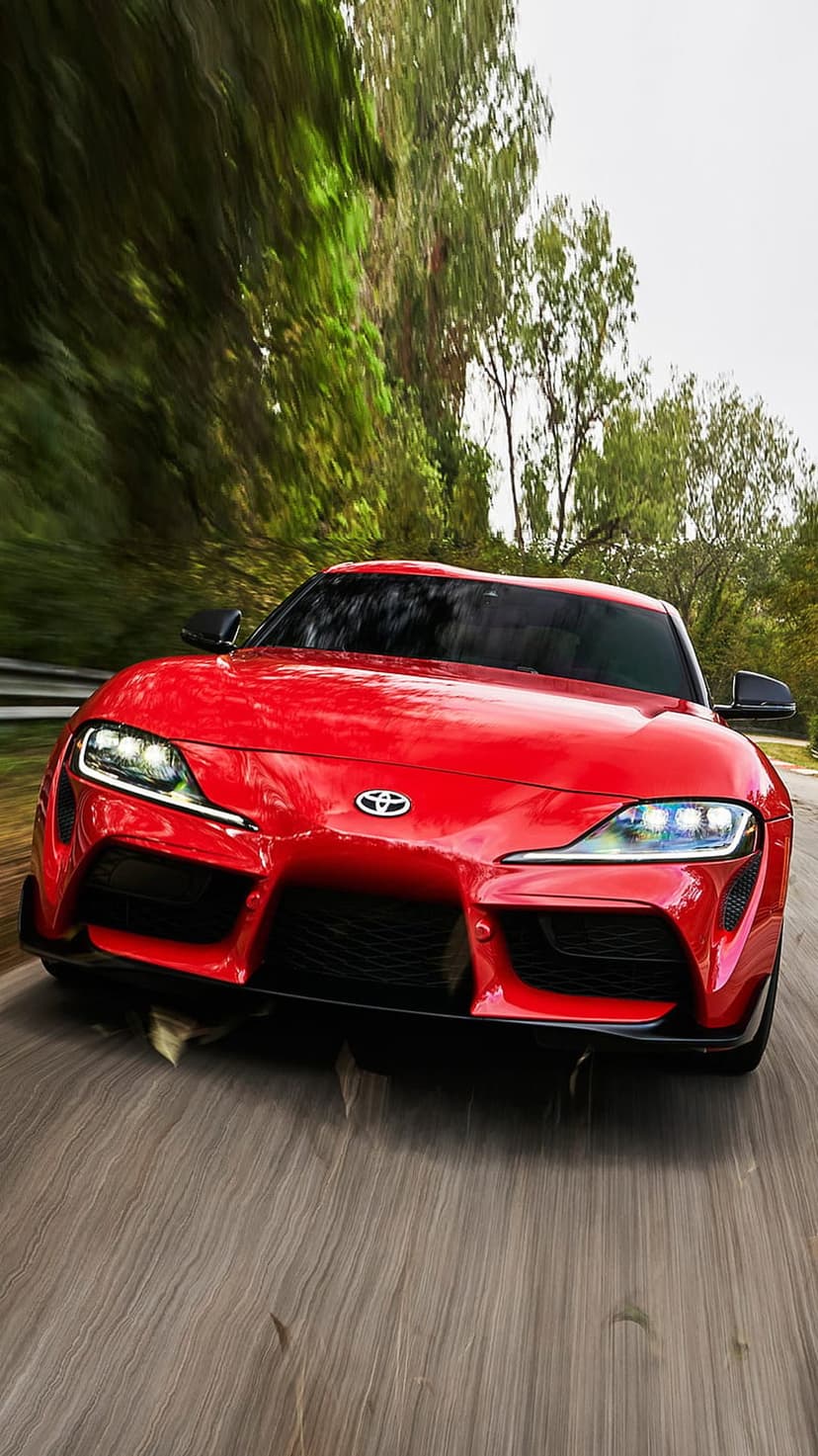 Red Toyota Supra car drives on a winding road with blurred green trees and sharp LED headlights visible.