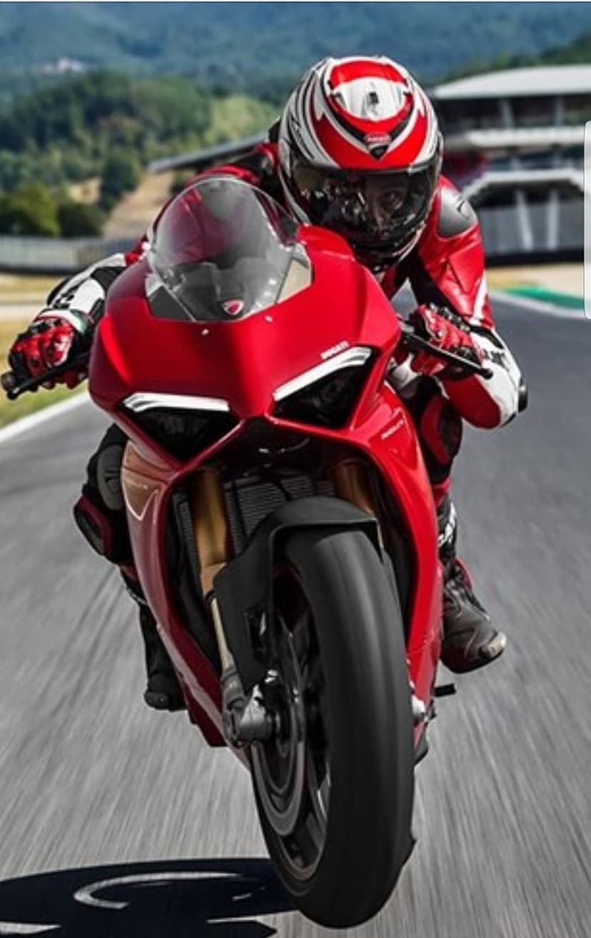 Ducati rider in red racing leathers leans a motorcycle into a sharp turn on a sunlit asphalt racetrack.