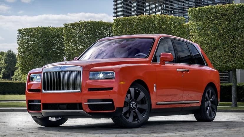 Orange Rolls-Royce Cullinan luxury SUV parked on a stone plaza with green hedges and a modern building behind it.