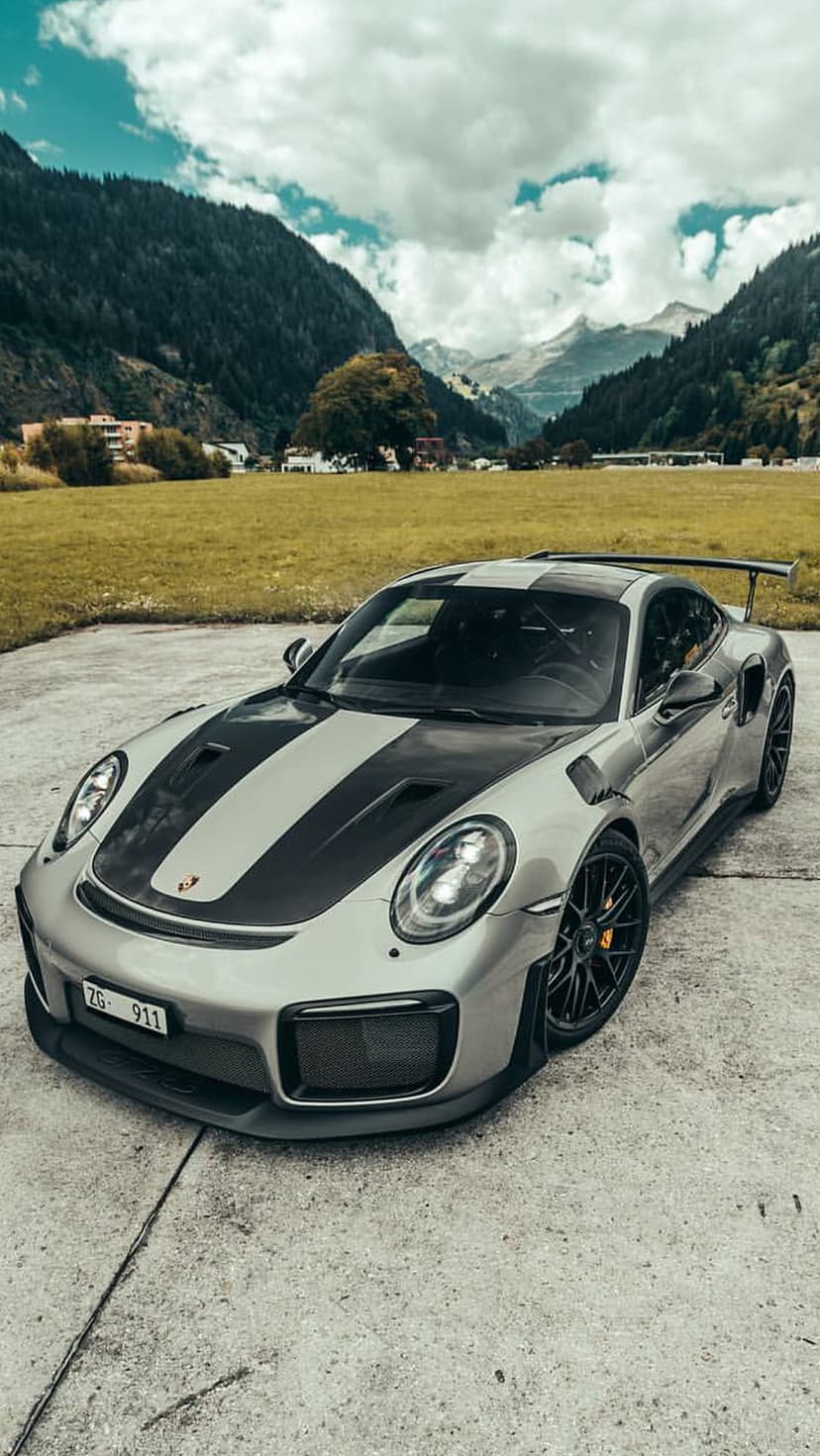 Silver Porsche 911 GT2 RS with black stripes parked on concrete before green mountains and a cloudy sky.