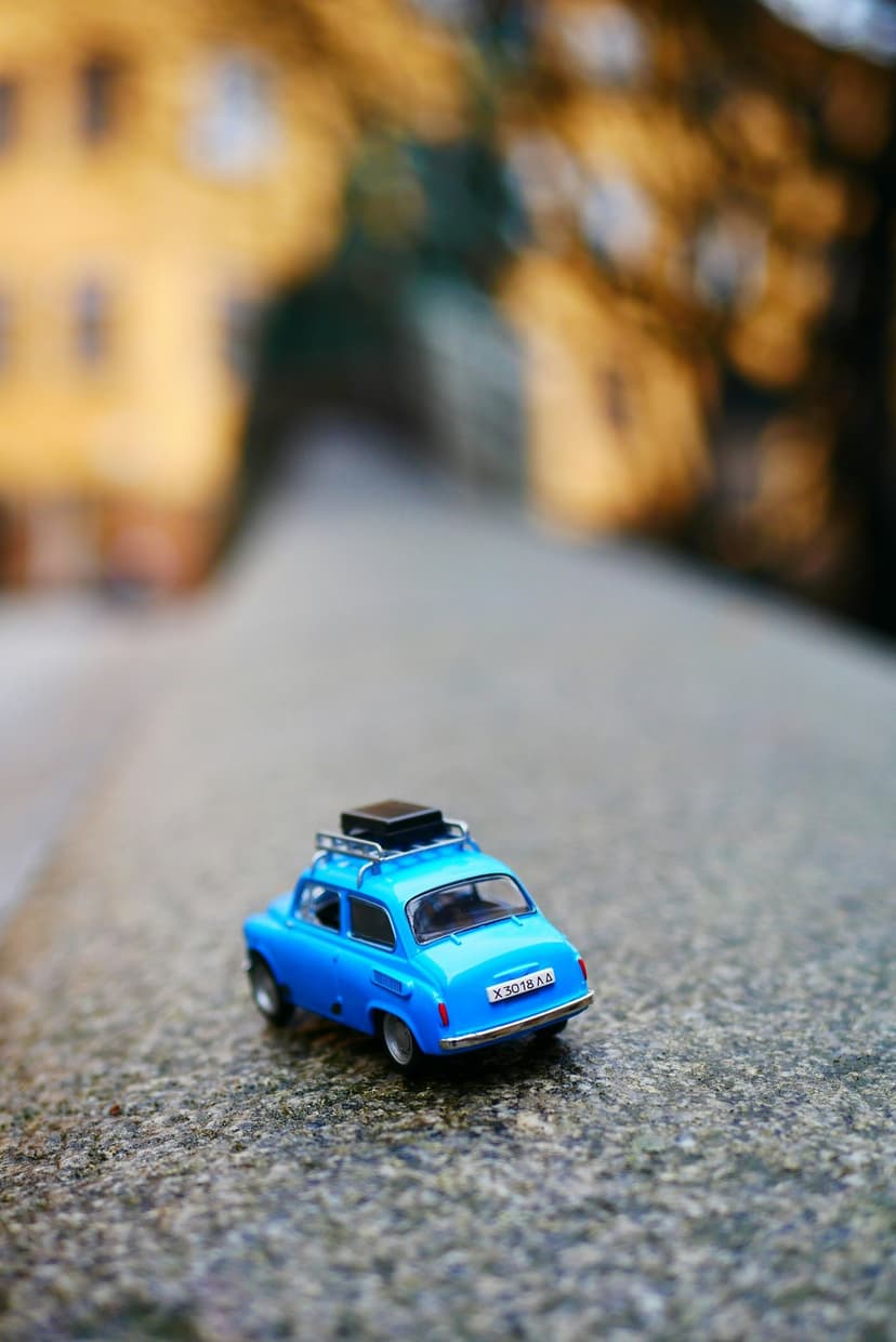 Miniature blue car with a luggage rack on a cobblestone path against a blurred yellow and green background.