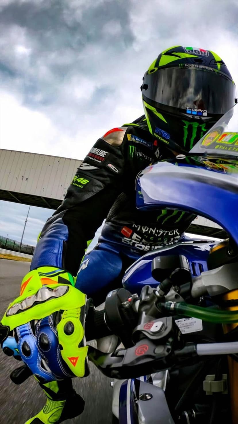 Motorcycle racer in a yellow and blue helmet grips the handlebars of a blue Yamaha sportbike on a racing circuit.