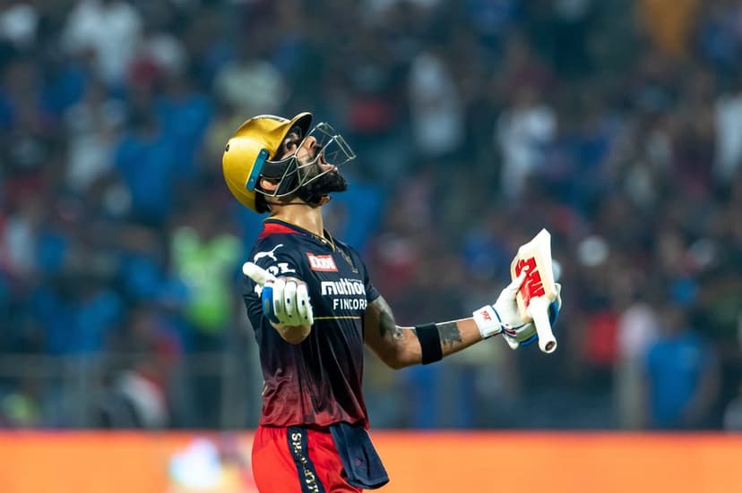 Virat Kohli raises his cricket bat and helmet in triumph during a night match with stadium lights in the background.