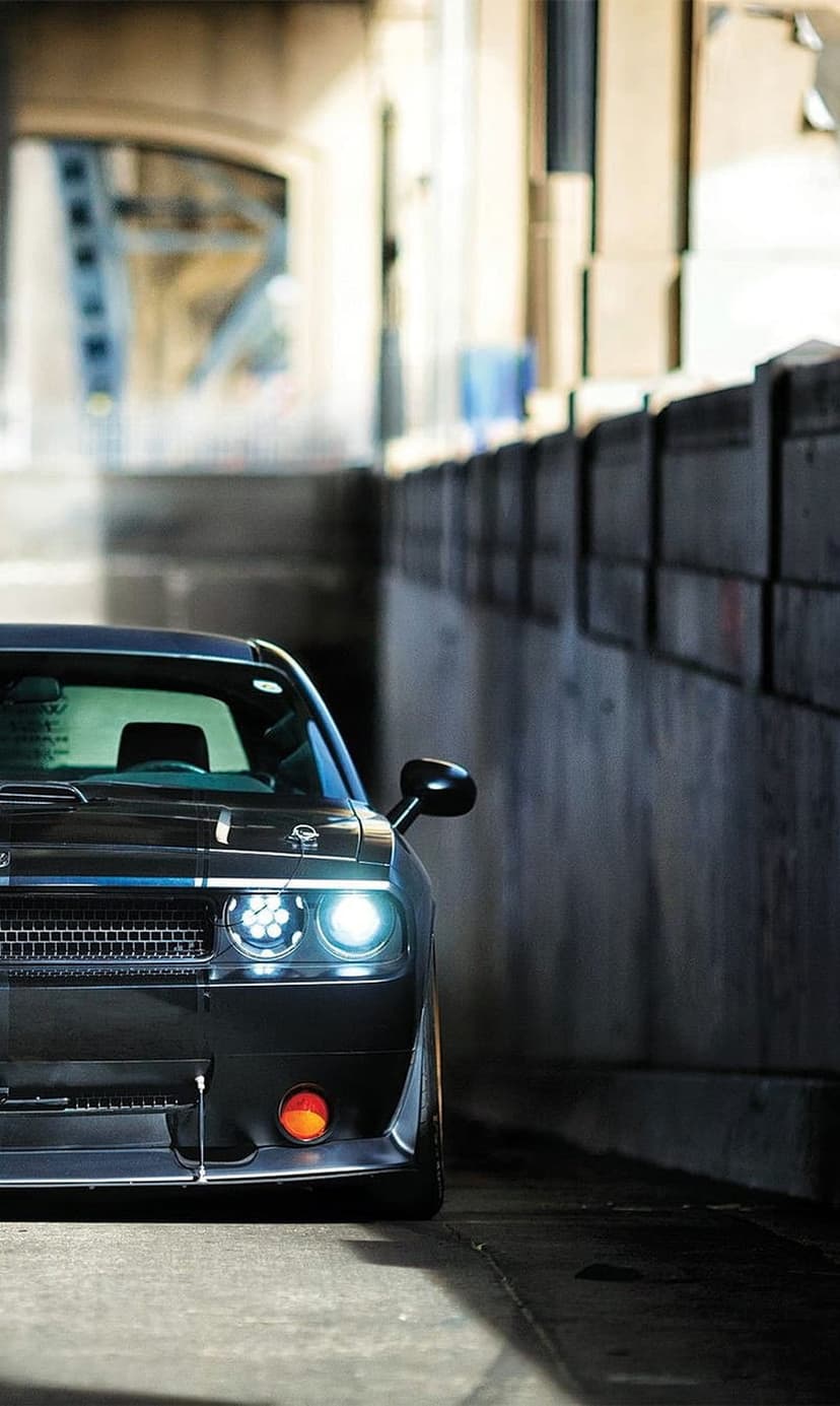 Dodge Challenger with glowing headlights parked in a dark concrete underpass beneath an architectural bridge.