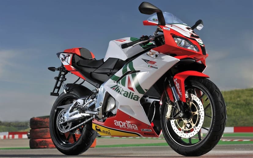 Aprilia sportbike in Alitalia racing livery parked on a sunny racetrack with a clear blue sky background.